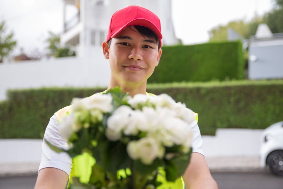 A delivery person wearing a red cap and yellow high-visibility vest holding a bouquet of fresh white flowers, with bright petals and green stems, arranged in a compact, rounded style. The bouquet is w