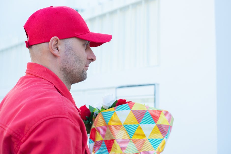 A delivery person dressed in a red cap and jacket is holding a brightly wrapped bouquet of flowers with a colorful geometric pattern, consisting of red roses, white blooms, and green foliage. The bouq