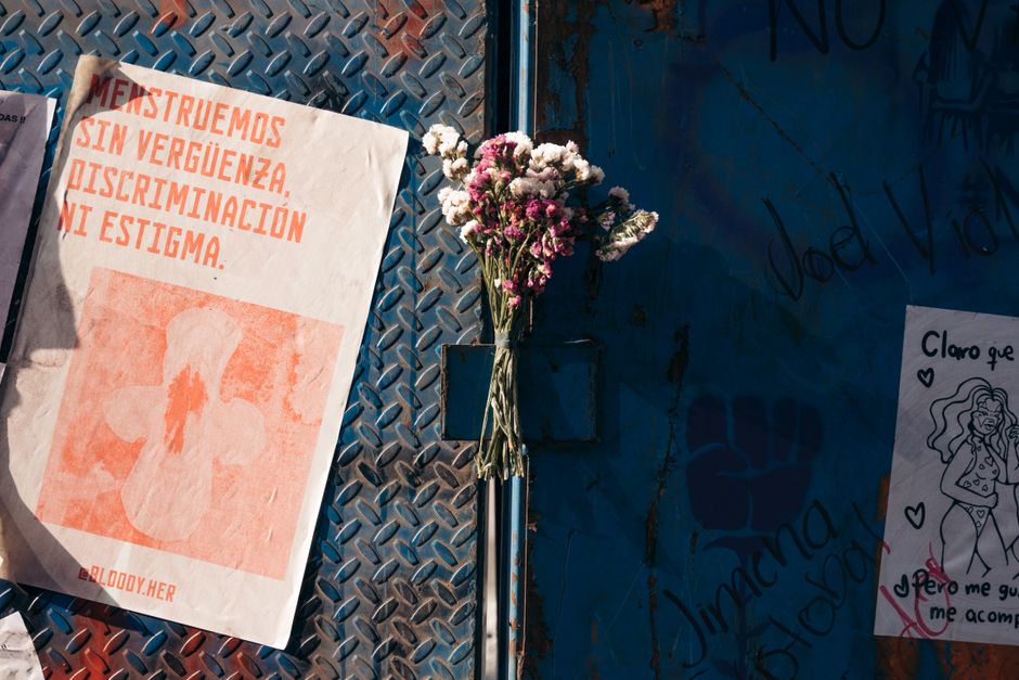 A bouquet of small pink and white carnations with ruffled petals and green stems, held together by a natural twine wrap, is positioned against a textured, blue-painted wall. To the left, a white poste