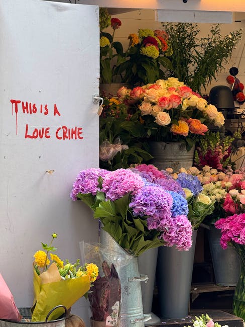 Inside a flower shop, a variety of fresh flowers including pink, purple, yellow, and white hydrangeas, roses, and other blooms are displayed in vases and buckets. The flowers are arranged on shelves a