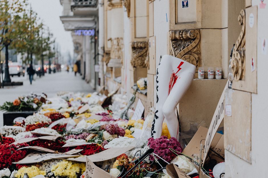 A sidewalk memorial display consisting of numerous floral tribute arrangements placed against a building wall, featuring a variety of fresh flowers including white chrysanthemums, red carnations, pink