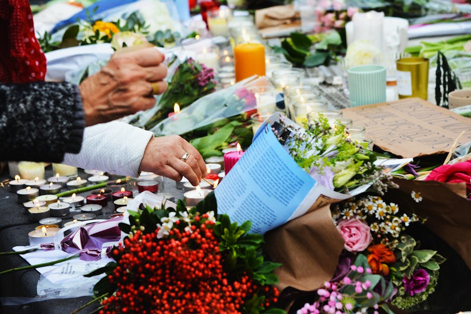 A somber floral memorial display featuring an array of fresh flowers, including pink roses, white daisies, and clusters of red berries, arranged on a table. The flowers are wrapped in brown kraft pape