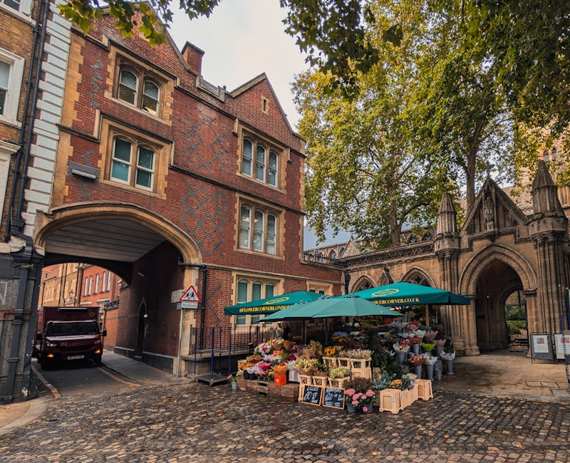 A flower stall set up on a cobblestone street under a green umbrella with the URL 'florimonovalondon.co.uk' printed on it, displaying a variety of fresh flowers including roses, carnations, and tulips
