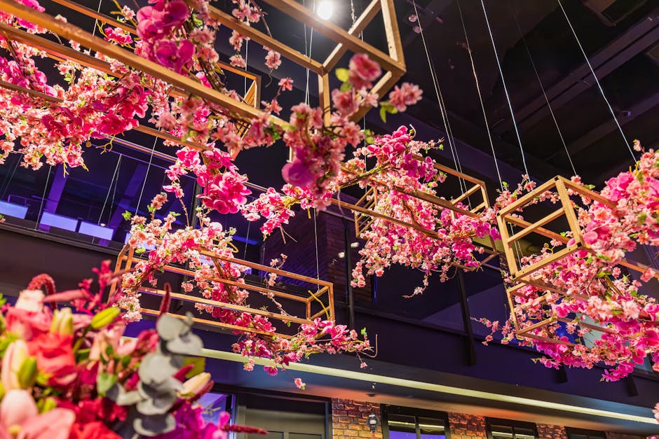 An elaborate floral installation featuring cascading pink cherry blossoms and other pink flowers suspended within gold geometric frames hanging from the ceiling of an indoor event space. The arrangeme