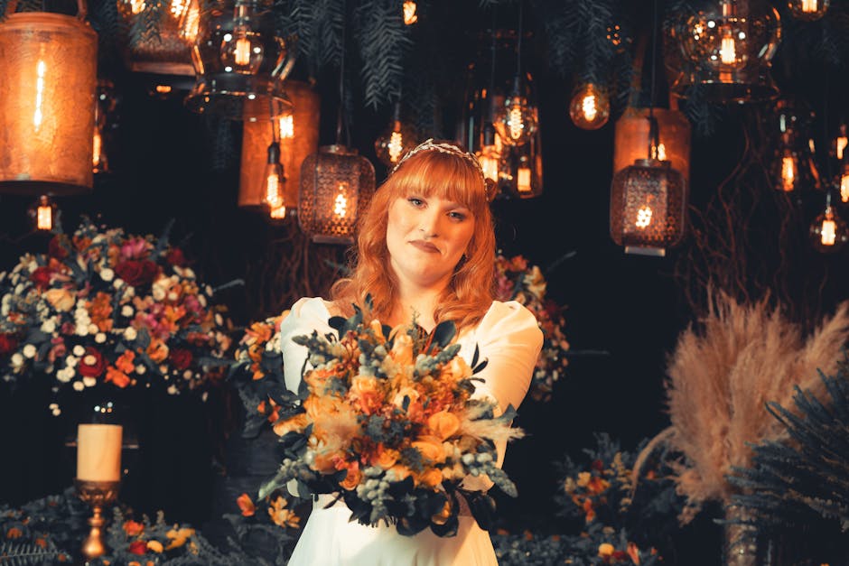 A woman with red hair and a gentle smile holds a large, colorful floral bouquet with a mix of orange, peach, and muted blue flowers, accented by greenery. She is dressed in white with puffed sleeves a