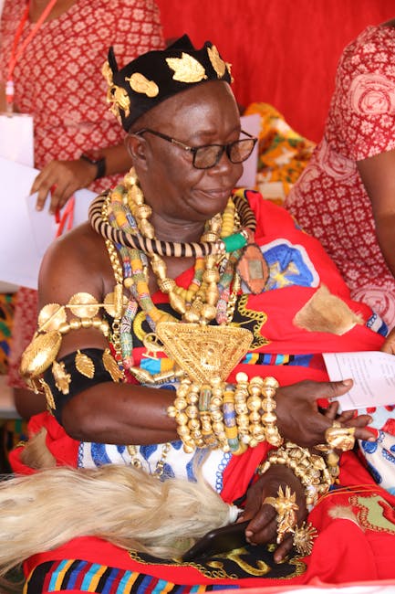 A woman dressed in traditional Maasai attire adorned with elaborate gold jewelry, including necklaces, bracelets, and rings, sitting on a red patterned fabric background. She wears a black headpiece d